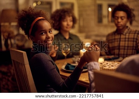 Happy African American girl holding hands with her family during Thanksgiving prayer at dining table and looking at camera.