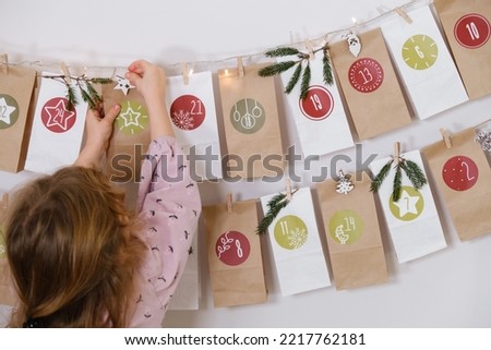 Toddler child gets new advent calendar task, waiting for Christmas. Festive mood for cute curly girl. Kid takes advent bag with dates from the wall at home.