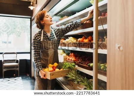 Caucasian young adult employee is working on adding the stock of fruit to the shelf before the grocery store opens. Saleswoman with an apron is holding fruit basket and adding bell peppers to shelf.