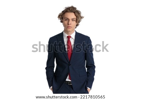 picture of young businessman with blue eyes and curly hair holding hands in pockets while wearing navy blue suit with red tie and posing on white background, portrait