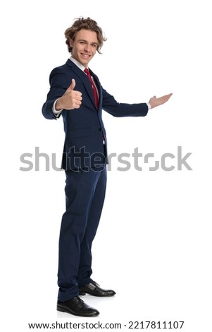 full length picture of smiling elegant businessman in suit making thumbs up gesture and showing to side, being happy and posing in a side view pose on white background