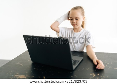 Tired blonde little girl putting broken hand wrapped in white plaster bandage on head thinking looking to laptop screen sitting at table on white background. Concept of child insurance and healthcare.