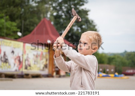 Little boy outdoors. The kid is holding a wooden ax. Boys playing in the street. Kids Games, carelessness, wooden toys - Concept of carefree childhood.