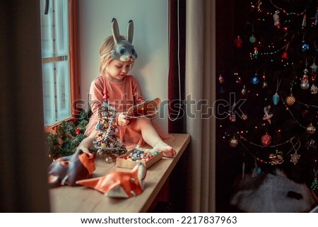 Cute little girl in mask is dressing up a tiny Christmas tree and trying on New Year's animal masks. Image with selective focus and toning. 