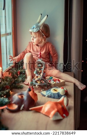 Cute little girl in hare mask is looking in snowy window, dressing up a tiny Christmas tree and trying on New Year's animal masks. Image with selective focus and toning. 