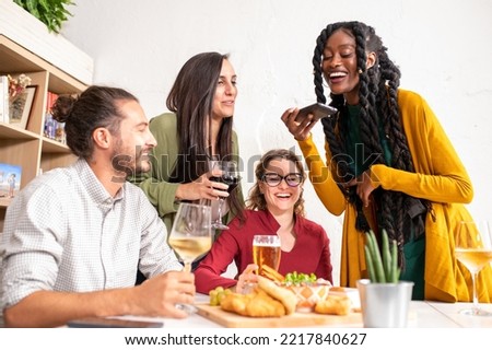 Young African woman smiling while sitting with her friends in a bistro, people taking photos of their meal with her smartphone, influencer enjoying food and drink