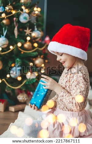 Smiling cute little girl in red Santa Claus hat opening a gift box sitting near Christmas tree. Merry Christmas and Happy New Year concept.