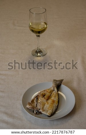 a white plate with a grilled fish and a glass of cold white wine on a table with a beige linen tablecloth