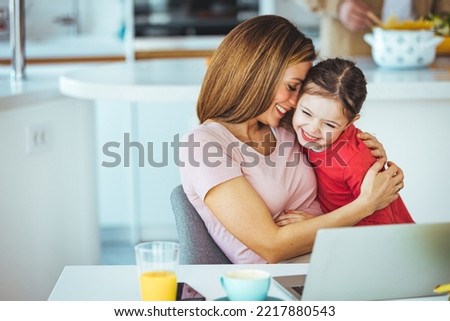 Mother and daughter using laptop and Internet. Freelancer workplace in cozy kitchen. Woman and child girl together. Concept of female business, working mom, freelance, home office. Lifestyle moment.