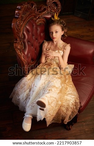 Fairy tales. Beautiful little princess girl in a golden dress and a crown sits in an armchair in her vintage castle. Full length shot.  Royalty-Free Stock Photo #2217903857