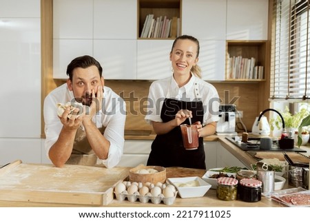 Handsome delighted husband looking at camera holding dough in arms and adorable young blonde wife with ponytail mixing tomato sauce in measuring cup with spoon. Baking cooking pizza.
