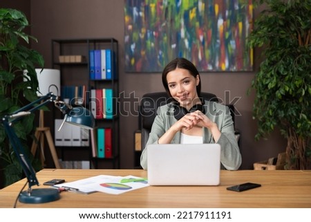 Photo of delighted young woman wearing headphones looking at camera smiling sitting at desk in front of computer laptop working studing process from home.