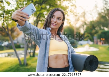 Beautiful african girl with fitness mat in sportswear standing in a park taking selfies after yoga practice. Outdoor lifestyle. Healthy active lifestyle