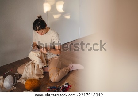 Focused young woman sewing in comforts of her apartment, embroidering a colourful picture on a loop. Cloth textile. Home decoration. Home design.
