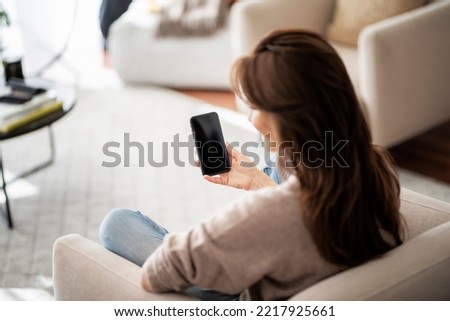 Rear view shot of an urecognizable woman holding smarpthone in her hand while relaxing at the armchair. Blank screen template mock up. Over the shoulder view. Royalty-Free Stock Photo #2217925661