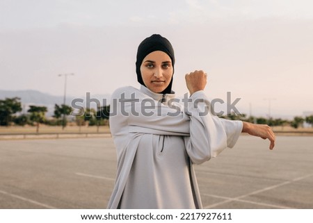 Beautiful Muslim woman looking at the camera while working out outdoors in the morning. Sporty woman with a hijab warming up by doing stretch exercises. Royalty-Free Stock Photo #2217927611