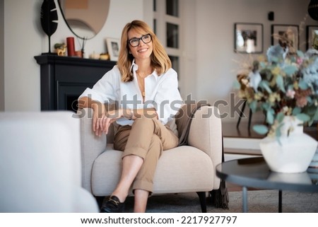 Portrait of attractive middle aged woman relaxing in an armchair at home. Blond haired female wearing eyeglasses and white shirt. Royalty-Free Stock Photo #2217927797