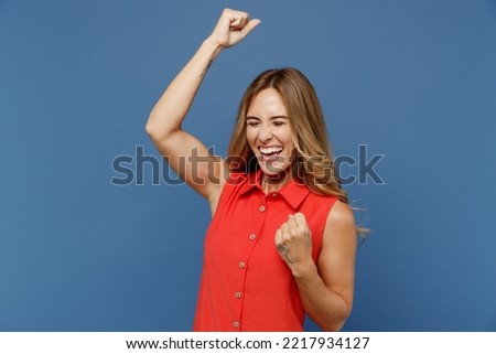Young excited caucasian woman 30s she wear red dress doing winner gesture celebrate clenching fists say yes isolated on plain dark royal navy blue background studio portrait. People lifestyle concept
