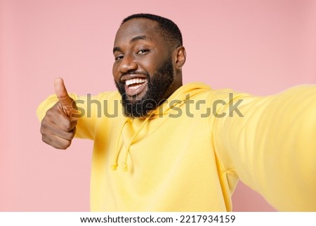 Close up cheerful young man of African American ethnicity 20s he wears casual basic yellow hoodie doing selfie shot pov on mobile cell phone showing thumb up isolated on pastel pink color background