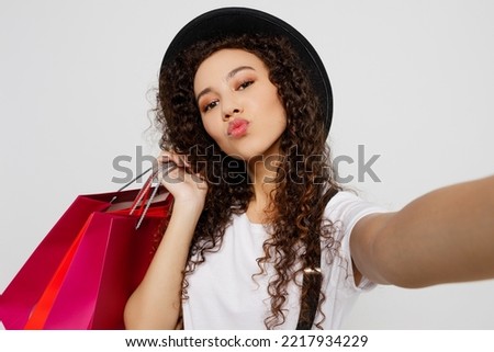 Close up young woman wears t-shirt hat hold in hand red paper package bag after shopping do selfie shot pov mobile cell phone isolated on plain solid white background Black Friday sale buy day concept