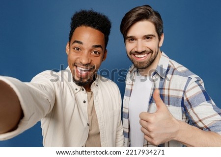 Close up young two friends happy men wear white casual shirts together do selfie shot pov on mobile cell phone show thumb up isolated plain dark royal navy blue background. People lifestyle concept