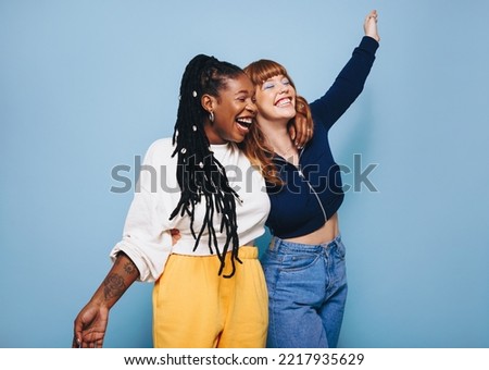 Two best friends laughing and having fun while embracing each other in a studio. Happy young women enjoying themselves while standing against a blue background. Friends making cheerful memories.