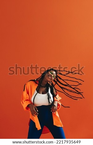 Happy young woman dancing and flipping her hair in a studio. Excited young woman celebrating and having fun against an orange background. Energetic young woman wearing dreadlocks and casual clothing.