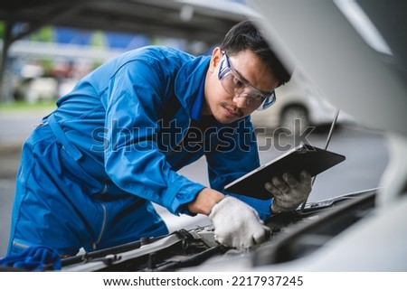 A young mechanic opens the hood of a car to check for engine damage and perform professional maintenance. He wears a blue uniform and inspects and repairs his car in the maintenance center. Royalty-Free Stock Photo #2217937245