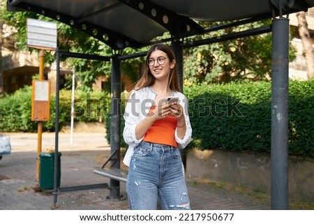 Smiling beautiful woman tourist holding mobile phone waiting for public transport on bus stop looking away, copy space 