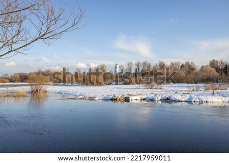 March sunny day by the river. A picturesque landscape, early spring, a river with snow-covered banks, dry grass and bushes. Church in the background. The first thaws, the snow is melting
