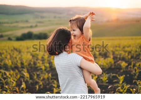 A young beautiful woman with her little baby daughter walking in the green corn-field on a sunny summer day at sunset. Parents and kids relationship. Summer