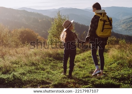 Traveler attractive young mother and pretty little daughter standing in grass on background beautiful calm autumn mountains at sunny day, enjoying nature. Lifestyle family travel tourism concept