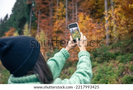 A young woman in a black hat takes a photo of the autumn forest on a smartphone.