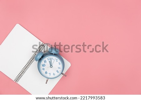 Alarm clock and empty notebook on a pink background, top view.