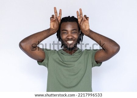 Portrait of happy African American man mimicking bunny ears. Cheerful young male model with braided dark hair in green T-shirt looking at camera, holding victory sign above head. Entertainment concept