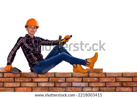 a young woman in a construction helmet with a construction hammer and a trowel in her hands is sitting on a brick wall isolated on a white background