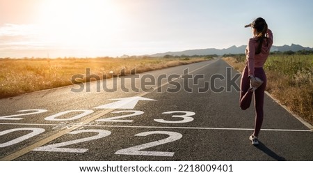 New year 2023 or start straight concept.word 2023 written on the asphalt road and athlete woman runner stretching leg preparing for new year at sunset.Concept of challenge or career path and change. Royalty-Free Stock Photo #2218009401