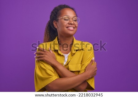 Young happy beautiful African American woman millennial with curly hair hugs herself to cheer up and get rid of loneliness dressed in casual yellow shirt, stands on isolated purple background. Royalty-Free Stock Photo #2218032945