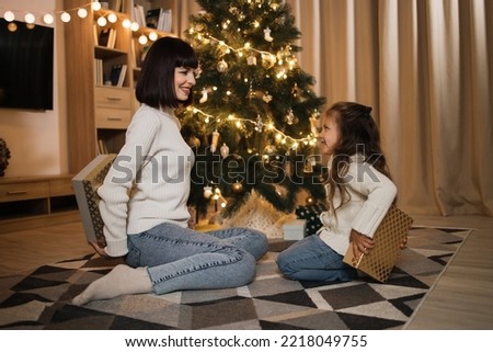 Attractive young caucasian mother and preschool little cute daughter girl sitting and holding boxes with gift near tree at evening at home. Merry Christmas.