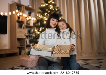 Focus on boxes with presents. Merry Christmas and Happy Holidays. Young happy mother and little cute daughter sitting with gifts after decorating tree in living room. Loving family indoors.