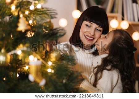 Happy small preschool caucasian child girl decorating Christmas tree with happy young mother, kissing mom in cheek enjoying preparing for New Year celebration at home, miracle time concept.