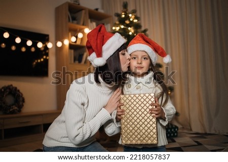 Attractive caucasian female sharing gift and kissing her cute preschool daughter in santa hat, sitting on floor on background of decorated tree, spending happy winter holidays at home.