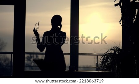 dark silhouette of Young business woman. she holds glasses in her hand, talks emotionally on mobile phone, gesticulates with hands, against background of large office window, at sunset, in rays of Royalty-Free Stock Photo #2218063719