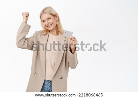 Beautiful smiling blond woman, senior lady showing credit card, recommending bank, standing in trench coat over white background