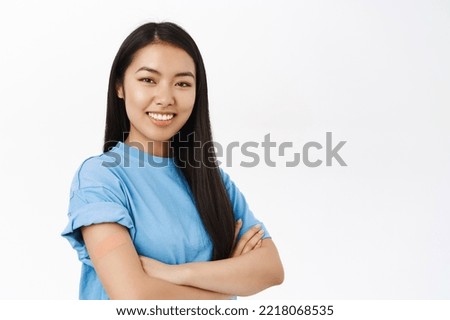 Covid-19, pandemic and healthcare. Smiling korean woman with band aid on shoulder, promotes vaccination, coronavirus vaccine, stands over white background