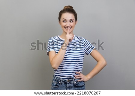 Portrait of woman wearing striped T-shirt showing shh gesture holding finger near lips, with naughty smile on face, making surprise, secret. Indoor studio shot isolated on gray background.