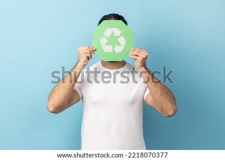 Portrait of anonymous man wearing white T-shirt hiding face behind green recycling sign, garbage sorting and environment protection. Indoor studio shot isolated on blue background. Royalty-Free Stock Photo #2218070377