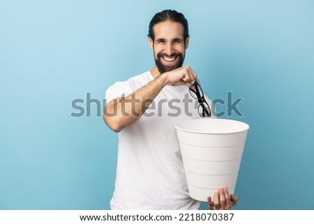Portrait of man with beard wearing white T-shirt throwing out eyeglasses into rubbish bin, treat his eyesight, looking smiling at camera. Indoor studio shot isolated on blue background. Royalty-Free Stock Photo #2218070387