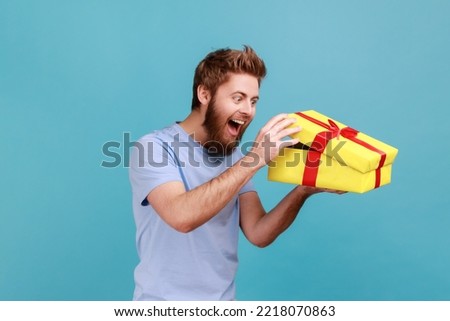 Portrait of excited handsome bearded man unboxing yellow gift box, looking inside with positive facial expression, birthday present. Indoor studio shot isolated on blue background. Royalty-Free Stock Photo #2218070863