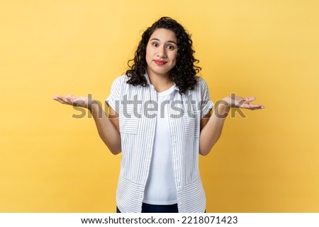 I am not sure. Portrait of ambiguous woman with dark wavy hair standing with raised arms, looking away and don't know what to do. Indoor studio shot isolated on yellow background. Royalty-Free Stock Photo #2218071423